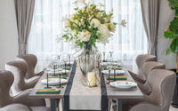 Belle table dressée, avec un grand bouquet de fleur au centre, des accessoires en métal avec des bougies, et un chemin de table blanc avec des liserés gris.