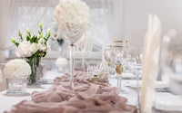 Chemin de table en tissu drapé beige rosé, sur une table avec une nappe blanche, décorée avec des fleurs blanches, des accessoires en verre et des bougies blanches.