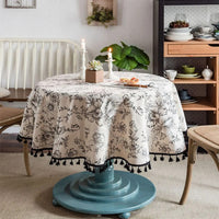 Nappe coton ronde et blanche avec motif floral, avec des pompons noir tout autour, dans une salle à manger, sur un table avec un pied bleu.